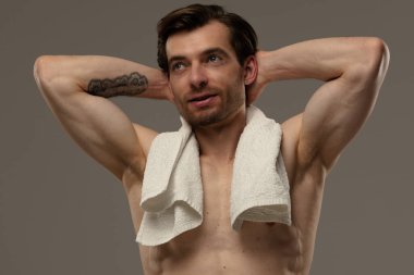 Üstsüz bir adam, omuzlarında havluyla, kendine güvenen bir şekilde poz veriyor.