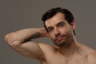 Rahatlamış ve kendinden emin bir adam kameraya gülümserken saçına dokunur. Sıradan ve dostane bir tavır sergiler..