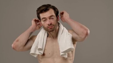 Üstsüz bir adam, omuzlarında havluyla kendine güvenen bir şekilde poz veriyor. Aydınlatıcı beyaz ışığın altında. Yavaş çekim