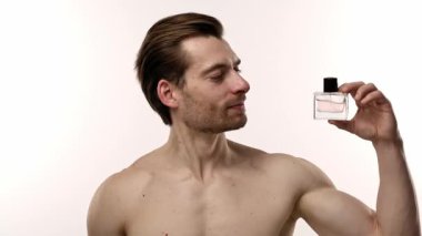 Beyaz arka planla işaretlenmiş, zarif ve sofistike bir şekilde parfüm şişesine bakan bir adam. Yavaş çekim