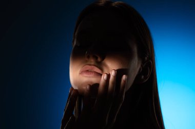 Yüzünde gölgeler olan bir kadının dingin portresi, canlı mavi bir arkaplan üzerine kurulmuş..