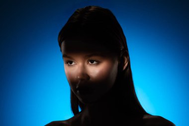 Yüzünde gölgeler olan bir kadının dingin portresi, canlı mavi bir arkaplan üzerine kurulmuş..