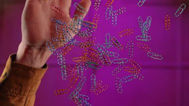 Havada süzülüyor gibi görünen çok renkli ataçların tuhaf bir görüntüsü, mor arka planda ellerle etkileşime giriyor. Yavaş çekim