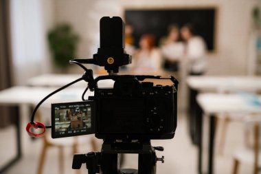 Sahne arkasında. Tripoddaki profesyonel bir video kamera, parlak ışıklandırılmış bir okul sınıfındaki bir grup sahnesini görüntüleyen bir ekrana odaklanır..