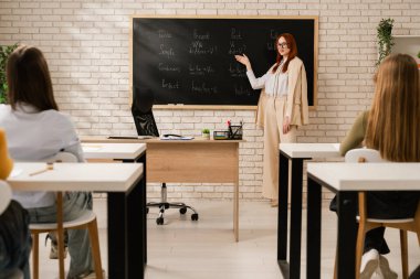 Bir öğretmen öğrencilerin önünde duruyor, modern bir sınıf ortamında tahtada İngilizce gramerini anlatıyor..