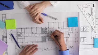 İşbirlikçi ekip, elleri masanın üstünde mimari planları inceliyor. Araçlar cetvel ve not defterlerini içerir. Üst çekim