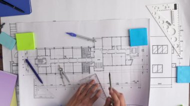 Üzerinde cetvel, pusula ve yapışkan notların yer aldığı, elleri ve çizim araçları olan ayrıntılı mimari plan. Üst çekim