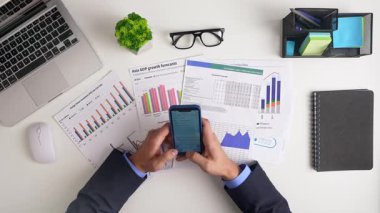 Bir iş adamı akıllı telefon ve dizüstü bilgisayarla grafikleri ve grafikleri inceleyerek ekonomik büyüme tahminlerini analiz eder. Üst çekim
