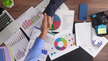 Resim, çeşitli finansal belgeler, tablolar ve grafikler üzerinde işbirliği ve mali analizi sembolize eden bir iş tokalaşmasını gösteriyor. Üst çekim, yavaş çekim.