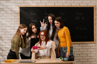 Enerjik öğrenciler öğretmenlerinin etrafında toplanıp selfie çekiyorlar, dostluk ve gayri resmi öğrenme sergiliyorlar. Arkaplandaki karatahta akademik bağlam ekler.