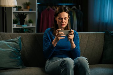 Bir kadın rahat bir şekilde kanepede oturuyor, üzgün ve akıllı telefonundan bir şeye bakıyor..