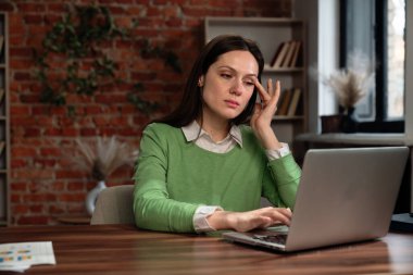 Yeşil tişörtlü bir kadın elinde dizüstü bilgisayarla masada oturuyor, rahat bir odada çalışırken duygusal görünüyor..