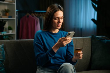 Kanepede oturan bir kadın elinde akıllı bir telefon ve kredi kartı ile online alışveriş yapıyor..