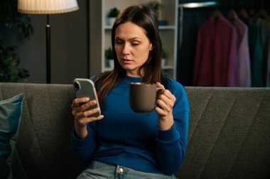 Bir kadın rahat bir şekilde kanepede oturur, akıllı telefonuyla uğraşırken kahvesini yudumlar, rahatlamanın tadını çıkarır..