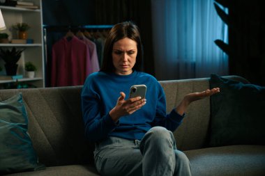 Mavi kazaklı, hayal kırıklığına uğramış bir kadın kanepeye oturur ve düşünceli bir şekilde telefonuna bakar ve kollarını açar. Bir elbise askısı ve bitkiler görünür..