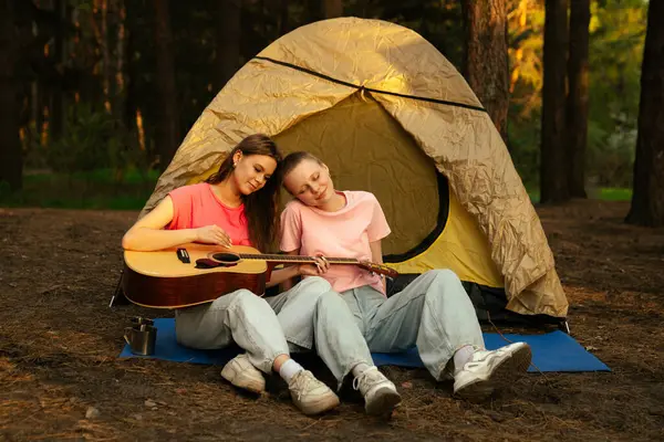 İki arkadaş ormanda bir çadırın yanında dinleniyor gitar çalıyor ve açık havada huzurlu bir ortam seyrediyor..