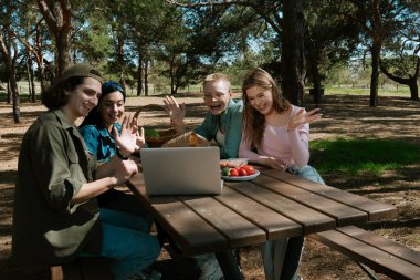 Bir grup arkadaş parkta bir piknik masasında yemeğin tadını çıkarırken bir yandan da bilgisayardan video görüşmesi yapıyorlar..