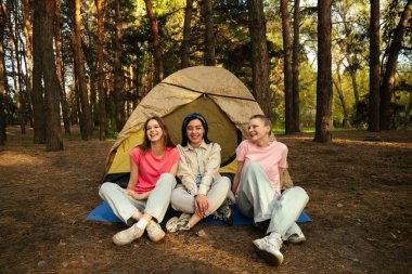 Üç kişi bir orman ortamında çadırın önünde mutlu bir şekilde oturur ve birlikte ferahlatıcı bir kamp deneyiminin tadını çıkarırlar..