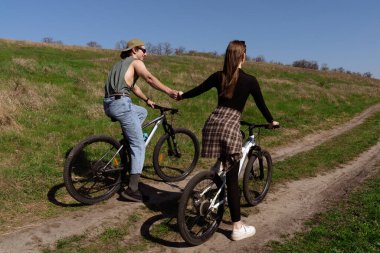 Kırsal bir yolda bisiklet süren, el ele tutuşan ve doğayla çevrili güneşli bir günün tadını çıkaran bir çift..