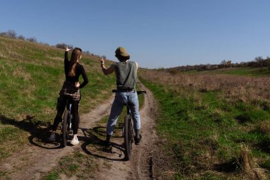 Güneşli bir günde yeşil çimenler ve açık mavi gökyüzüyle çevrili bir yolda iki kişi bisiklet sürüyor..
