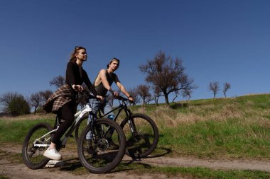 Ağaçlar ve mavi gökyüzüyle çevrili, temiz havanın tadını çıkaran iki kişi açık havada bisiklet sürüyor..