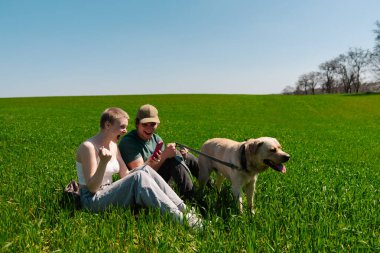 Neşeli bir çift yemyeşil çimen tarlasında oturuyor, güneşli bir günün tadını çıkarırken akıllı telefonları ve köpekleriyle meşgul oluyorlar..