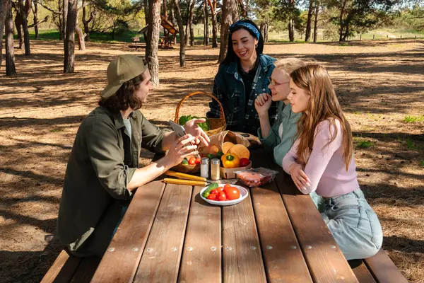 Bir grup arkadaş güneşli bir ormanda piknik yapıyorlar, sohbet ediyorlar ve açık havada taze sebzelerin tadını çıkarıyorlar..
