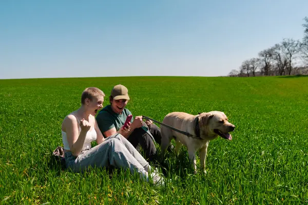 Neşeli bir çift yemyeşil çimen tarlasında oturuyor, güneşli bir günün tadını çıkarırken akıllı telefonları ve köpekleriyle meşgul oluyorlar..