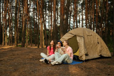 Üç arkadaş kamp alanlarında dinleniyor, manzaralı bir ormanda çadırın yanında sıcak içeceklerin tadını çıkarıyorlar..