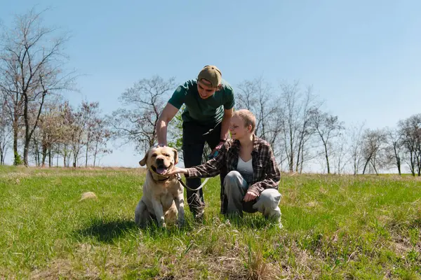 Güneşli bir günde arka planda ağaçlarla dolu yemyeşil bir arazide iki kişinin eğlendiği neşeli bir köpek sahnesi..