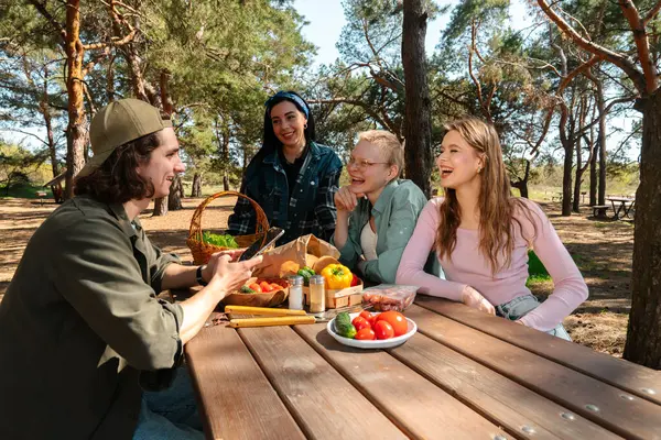 Bir grup arkadaş ormanda bir piknik masasında oturmuş taze sebzelerle yemek hazırlıyor ve birlikte gülüyorlar..