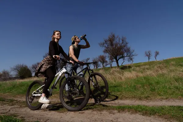 İki genç insan güneşli, çimenli bir arazide coşkuyla bisiklet sürüyor, açık hava aktivitelerine ve hareketli bir yaşam tarzına olan sevgilerini somutlaştırıyorlar..