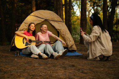 Arkadaşlarla dolu canlı bir kamp sahnesi müzik ve kahkahaların tadını çıkaran yemyeşil bir ormanda doğa ile çevrili..