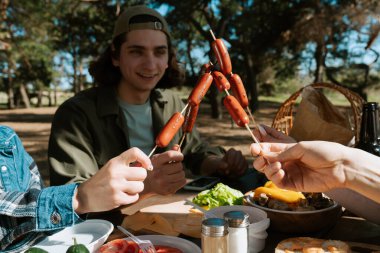 Bir grup park ortamında pikniğin tadını çıkarıyor. Dostça bir atmosferde ızgara sosis ve taze yiyecek paylaşıyorlar..