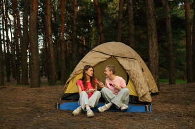 İki arkadaş çadırın yanında oturur, sohbet eder ve akıllı telefonlarıyla oyun oynarlar, ormanda yürüyüş yaparlar..