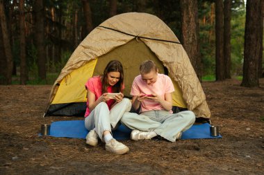 İki arkadaş çadırın yanında oturur, sohbet eder ve akıllı telefonlarıyla oyun oynarlar, ormanda yürüyüş yaparlar..