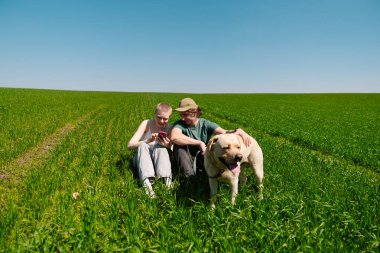 İki arkadaş yemyeşil bir alanda mutlu bir köpekle oturuyorlar. Berrak, mavi gökyüzünün altında birbirlerine eşlik ederek eğleniyorlar..