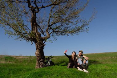 Genç bir çift yeşil bir tarlada bir ağacın altında otururken selfie çekiyor, açık gökyüzünün altında bisiklet sürüyor..