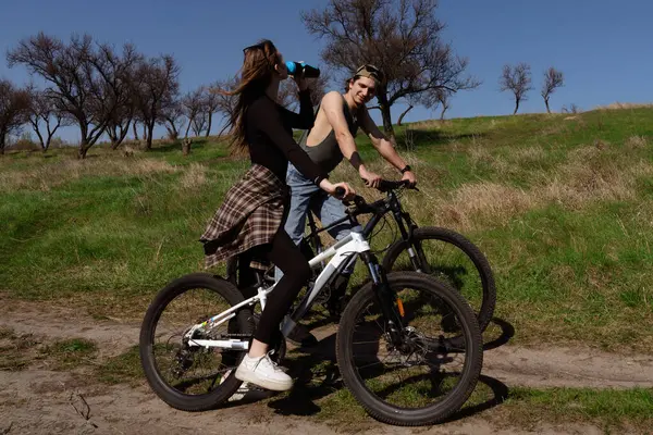 Güneşli bir günde parkta bir adam ve bir kadın dağ bisikleti sürüyorlar. Kadın şişeden içiyor..