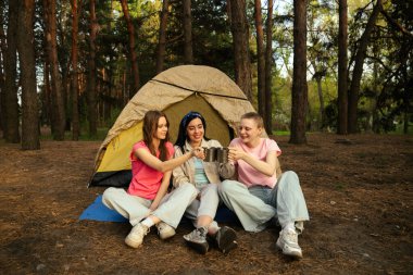 Üç genç arkadaş, huzurlu bir orman ortamının etrafında çadırın önünde otururken bir bardak içkiyi paylaşıyorlar..