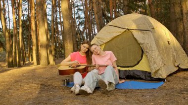 İki arkadaş ormanda bir çadırın yanında dinleniyor gitar çalıyor ve açık havada huzurlu bir ortam seyrediyor..