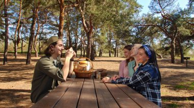Bir grup arkadaş ormanda pikniğin tadını çıkarıp anı fotoğrafla yakalıyorlar. Doğayla iç içe olup, bir piknik sepetiyle ahşap bir masanın etrafında toplanırlar..
