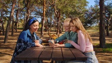Bir grup arkadaş, güneşli bir orman bölgesindeki ahşap piknik masasında otururken kahkaha ve atıştırmalıkların keyfini çıkarıyorlar..