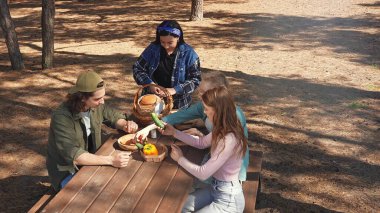 Bir grup arkadaş güneşli bir ormanda piknik yapıyorlar, sohbet ediyorlar ve açık havada taze sebzelerin tadını çıkarıyorlar..
