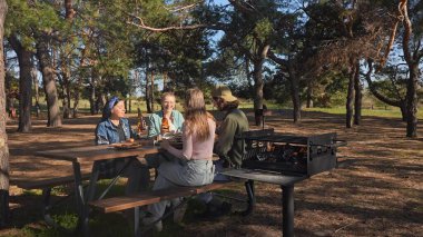 Bir grup arkadaş güneşli bir ormanda pikniğin tadını çıkarıyorlar. Önlerinde yiyecek ve içeceklerle ahşap bir masada oturuyorlar, neşeli ve rahat bir açık hava yaratıyorlar..