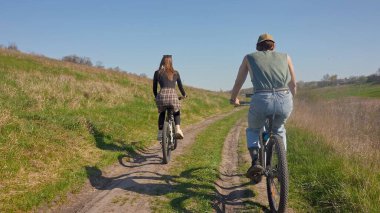 İki insan, açık mavi gökyüzünün altında yeşil ve açık alanlarla çevrili manzaralı bir toprak yolda bisiklet sürüyor. Arka plan