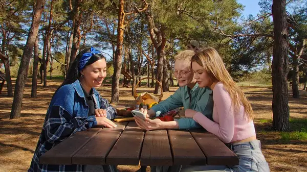 Bir grup arkadaş, güneşli bir orman bölgesindeki ahşap piknik masasında otururken kahkaha ve atıştırmalıkların keyfini çıkarıyorlar..