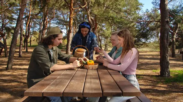 Bir grup arkadaş güneşli bir ormanda piknik yapıyorlar, sohbet ediyorlar ve açık havada taze sebzelerin tadını çıkarıyorlar..