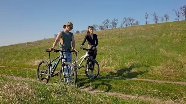 Ağaçlar ve mavi gökyüzüyle çevrili, temiz havanın tadını çıkaran iki kişi açık havada bisiklet sürüyor..