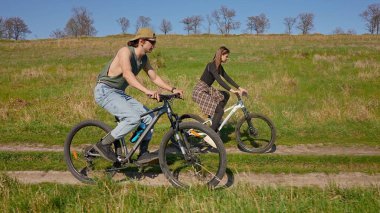 İki insan, açık mavi gökyüzünün altında yeşil ve açık alanlarla çevrili manzaralı bir toprak yolda bisiklet sürüyor. Yan görünüm